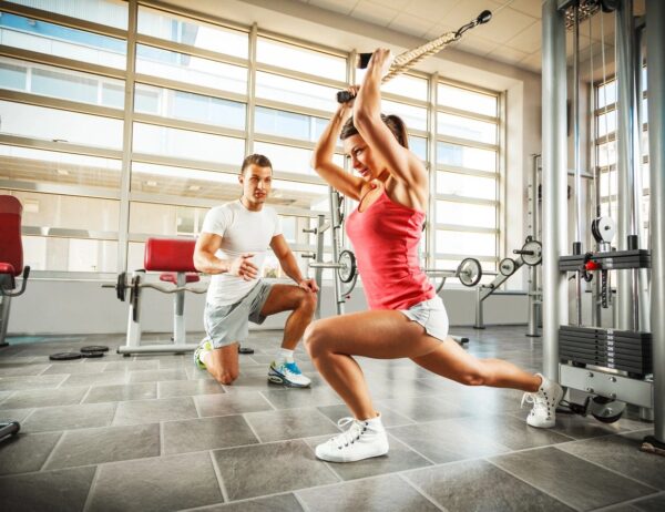 A woman is squatting in the gym while another man watches.